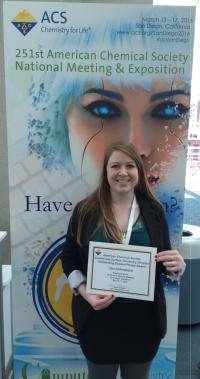 A student poses in front of a poster holding a certificate at Spring 2016 ACS Meeting