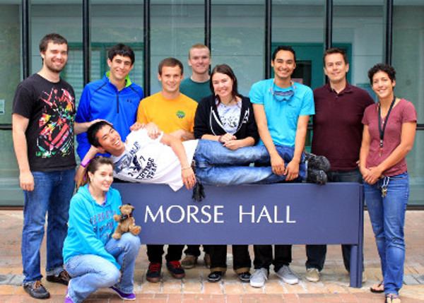 A group of students stand around the Morse Hall sign. A single student is lounging across the sign, his head supported by his hand.