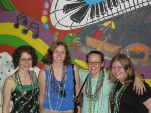 four women pose in front of a colorful wall depicting musical instruments and notes
